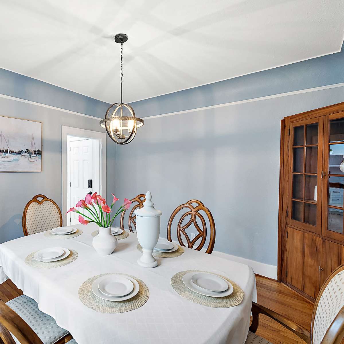 A dining room features a round table set with plates and a vase of tulips, surrounded by wooden chairs, with a cabinet and a painting on the walls.