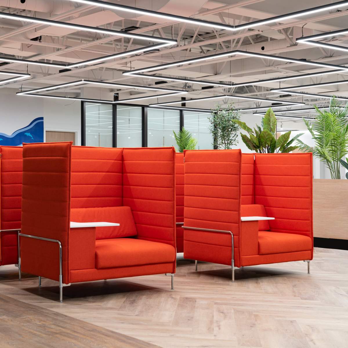 A modern office space featuring orange and gray seating pods with attached side tables, surrounded by indoor plants and decorative wall art.