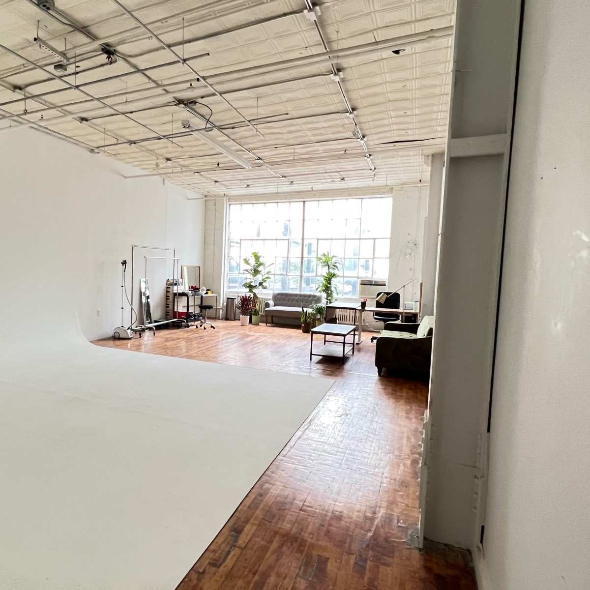 A spacious photography studio with a white backdrop and wooden flooring, featuring a seating area with green couches and large windows allowing natural light to enter.