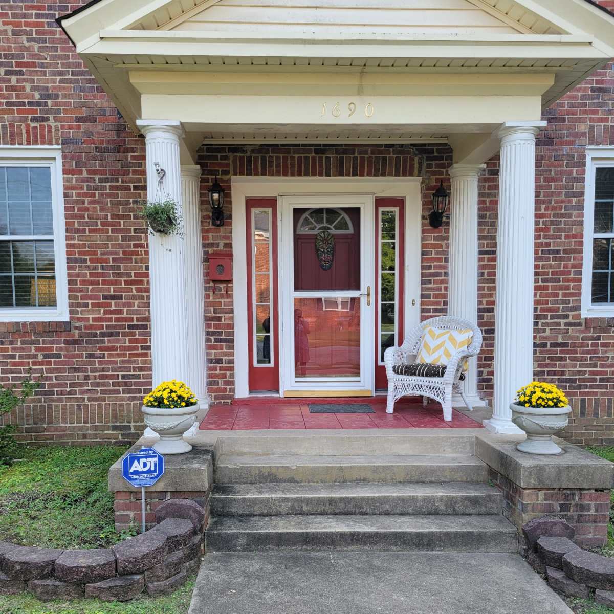A front entrance of a house with brick siding, featuring a red door, white columns, and potted yellow flowers on either side of the steps.