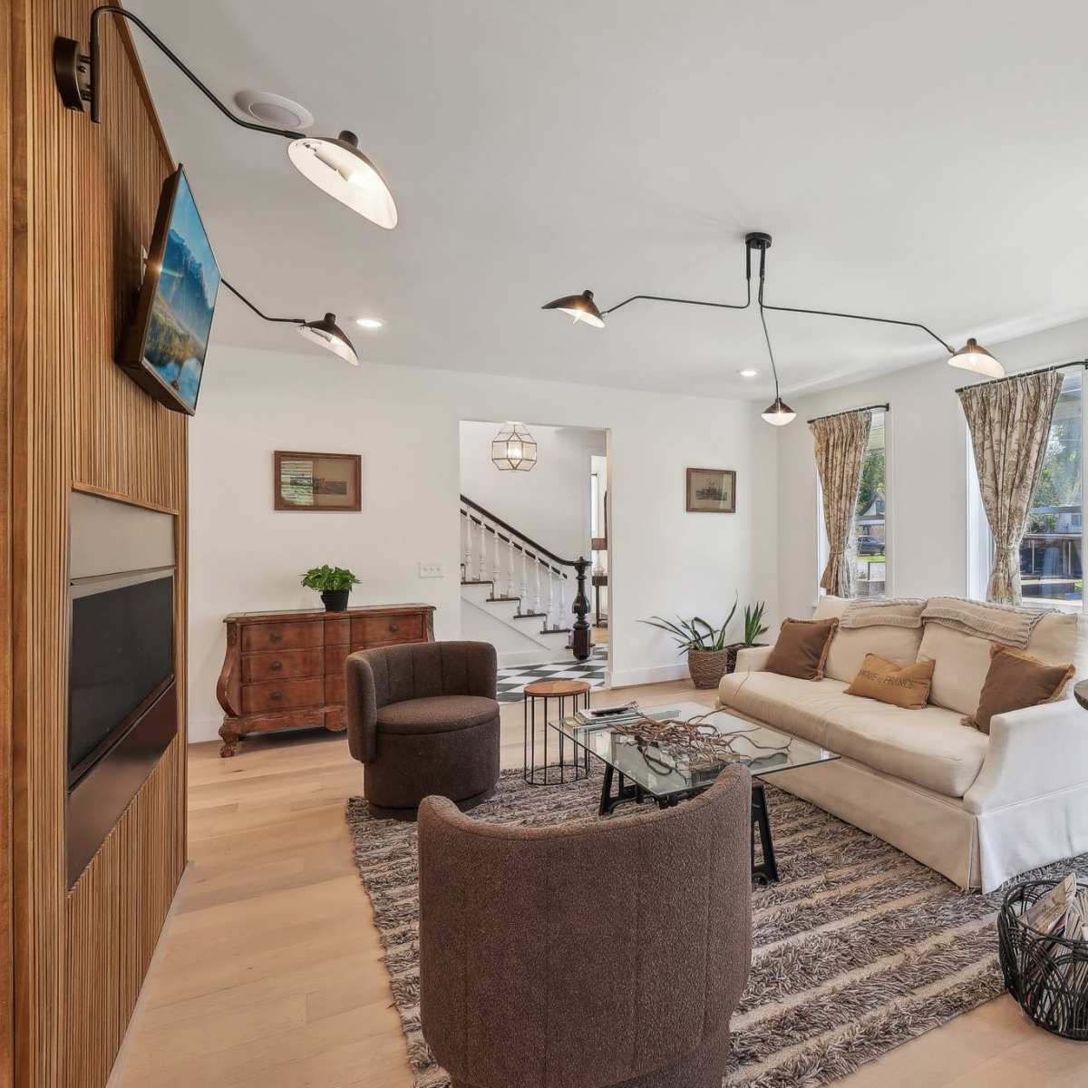 The living room features a light-colored sofa, two brown chairs, a glass coffee table, and patterned curtains, arranged around a central area with a decorative rug.