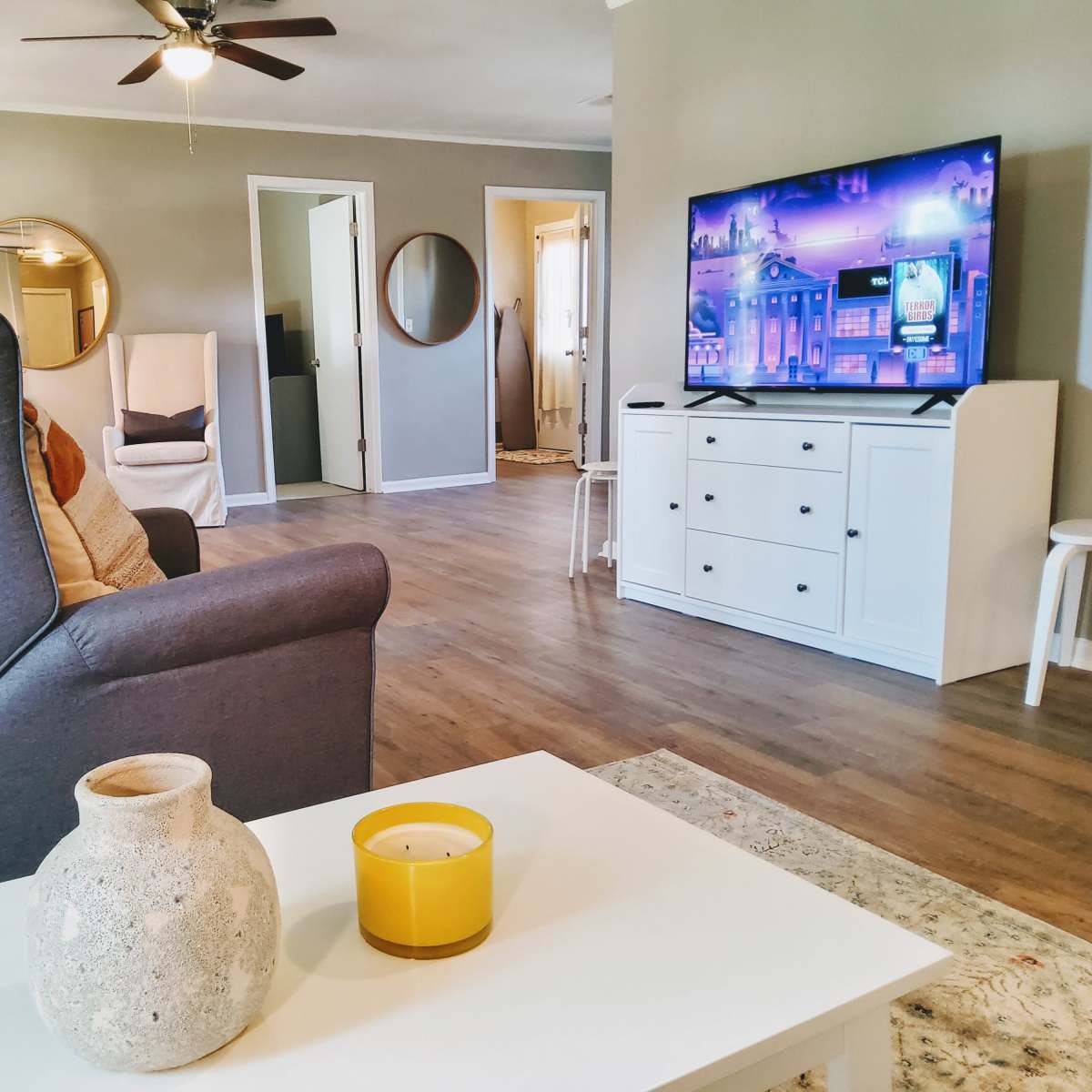 A living room featuring a grey armchair, a coffee table with a candle, a television against the wall, and a door leading to another room.