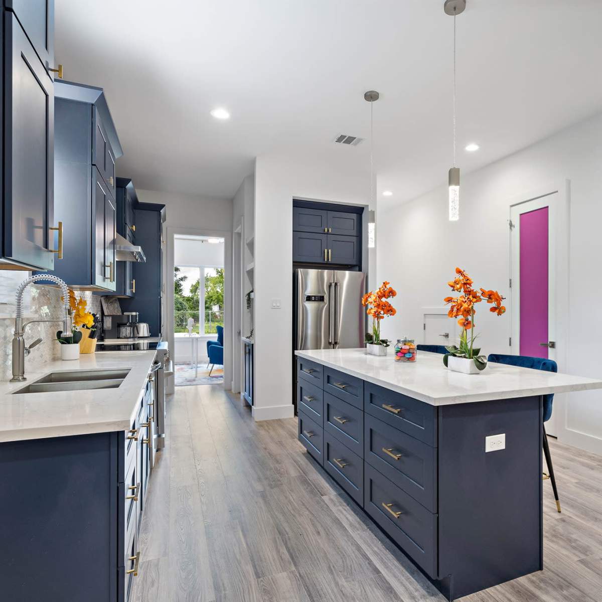 A modern kitchen with dark blue cabinetry, a central island with a white countertop, and a light-filled space featuring vibrant purple doors.