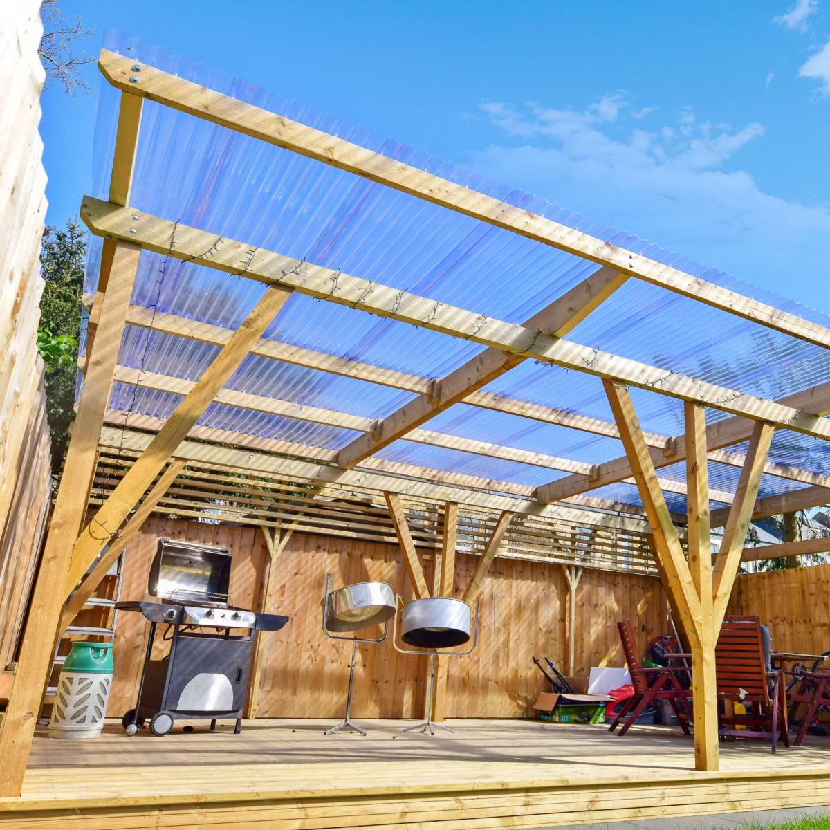 A wooden patio structure with a transparent roof, equipped with a barbecue grill and outdoor seating, situated in a backyard surrounded by wooden fencing.