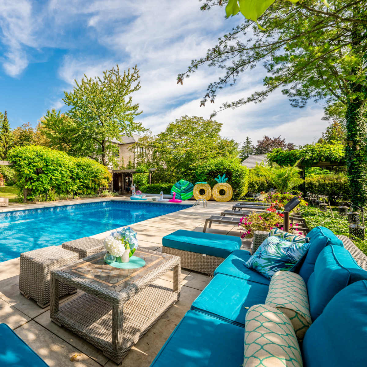 A backyard swimming pool surrounded by lounge seating and greenery, with a patio and dining area visible in the background.