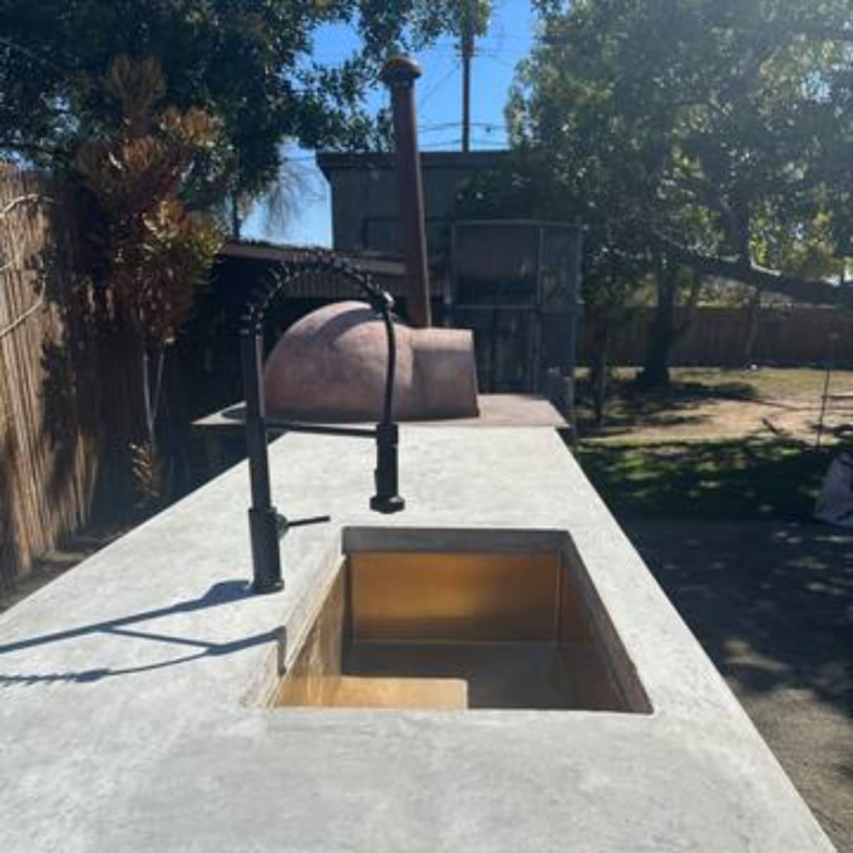 A concrete outdoor countertop with a sink and faucet, set in a backyard with a large tree and a building in the background.