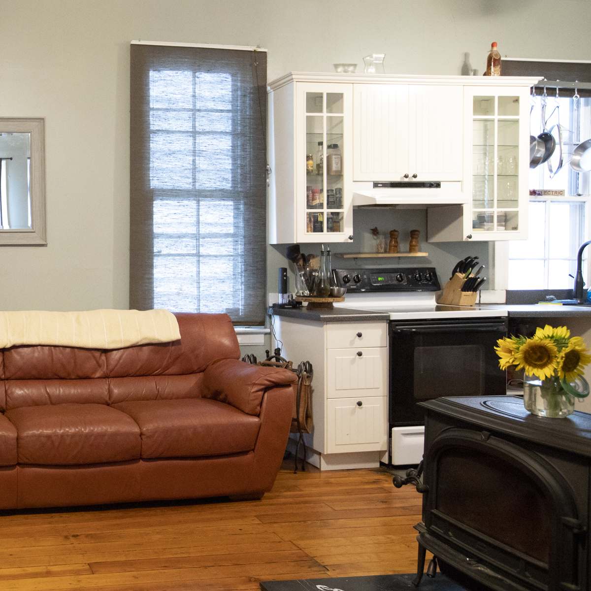 A living room with a brown leather couch, a small coffee table with sunflowers, and a modern kitchen area visible in the background.