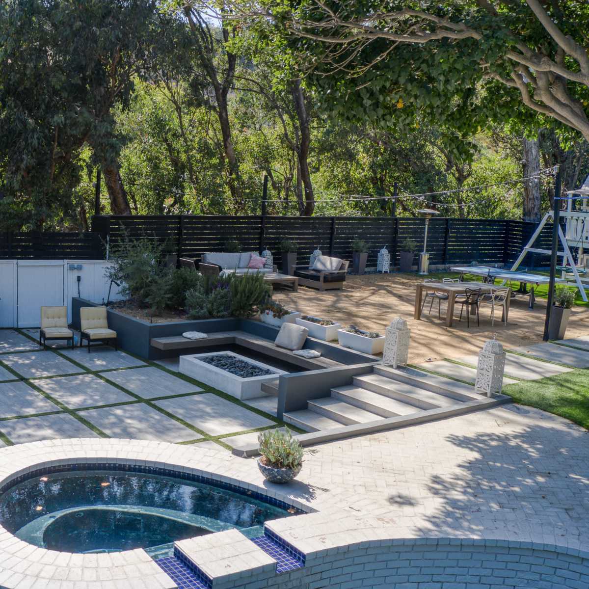 An outdoor patio area featuring a swimming pool, a bar with stools, a fire pit, and a lounge space surrounded by trees.