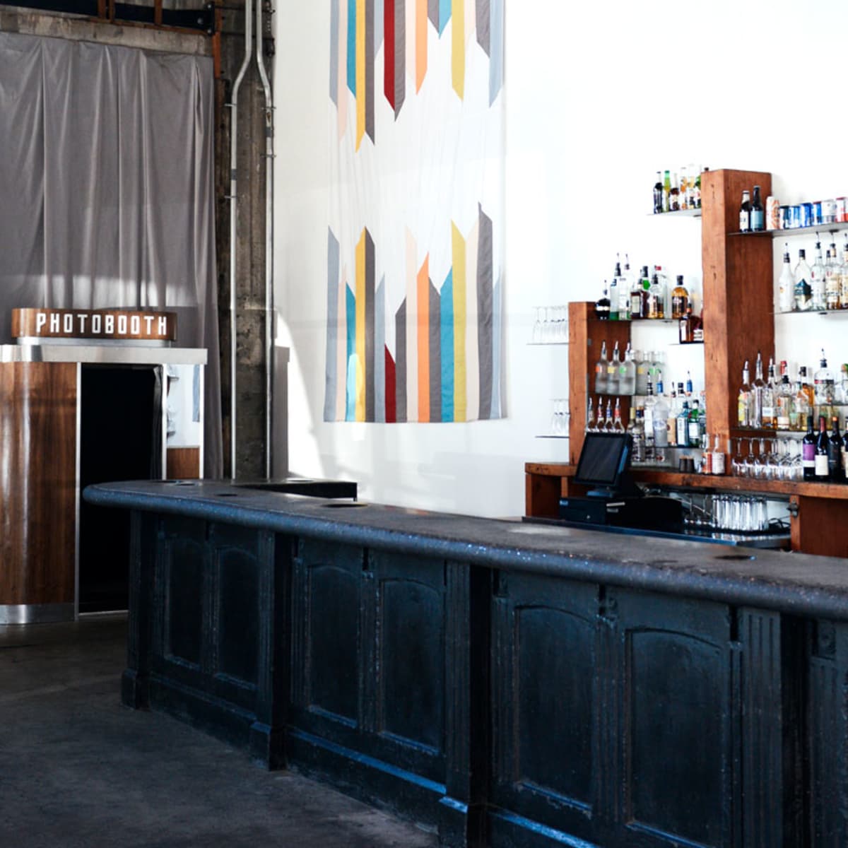 A bar area with a long, dark countertop, a shelf filled with various bottles in the background, and a photo booth to the left.