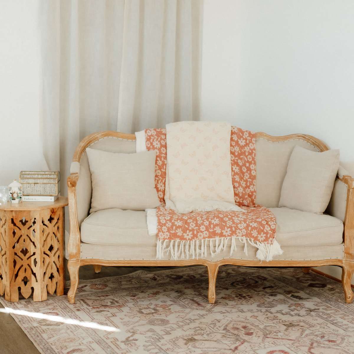 A vintage-style wooden sofa with light cushions is placed next to a carved wooden side table and a patterned rug.