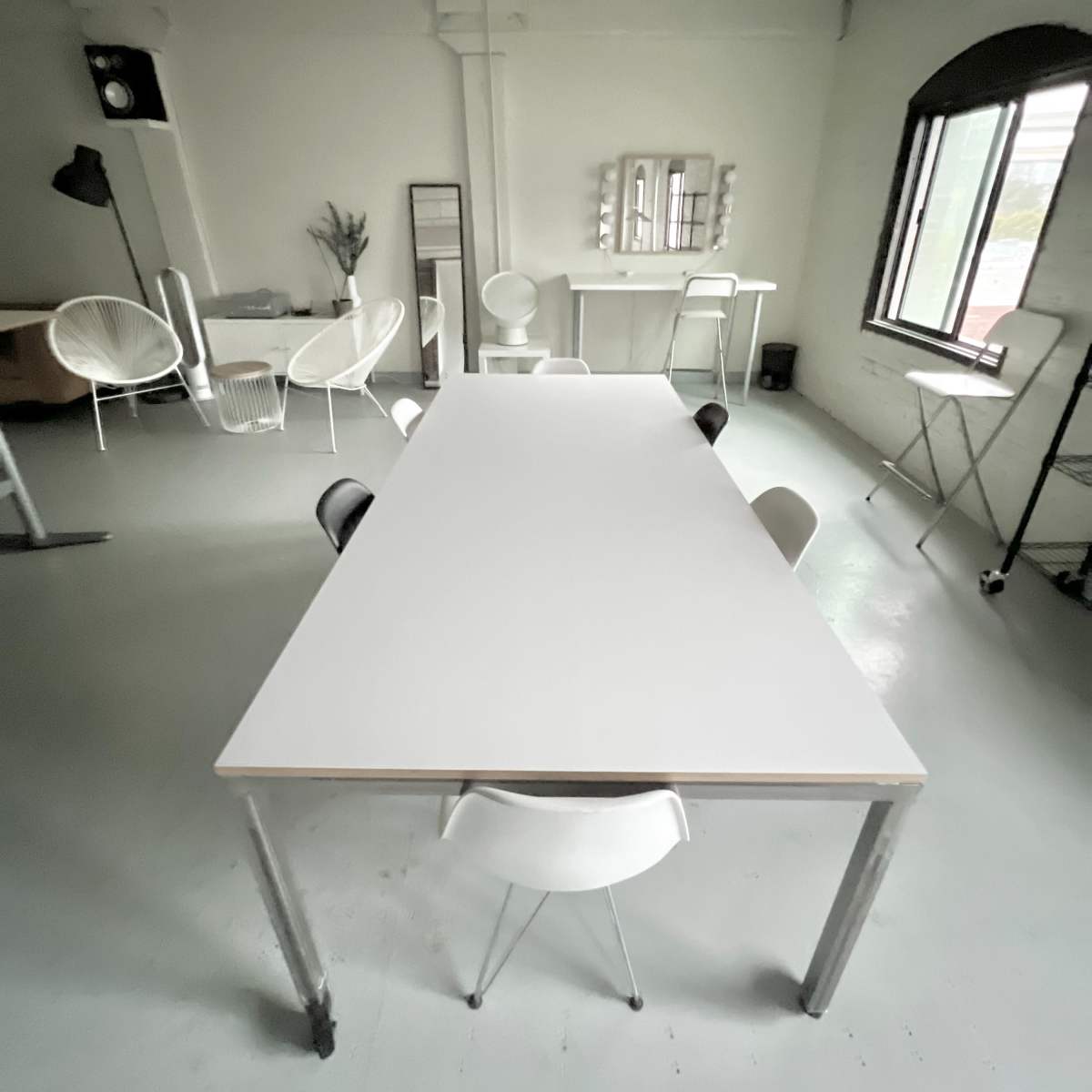 A spacious, minimalist room features a long white table surrounded by chairs, with additional furniture and large windows in the background.
