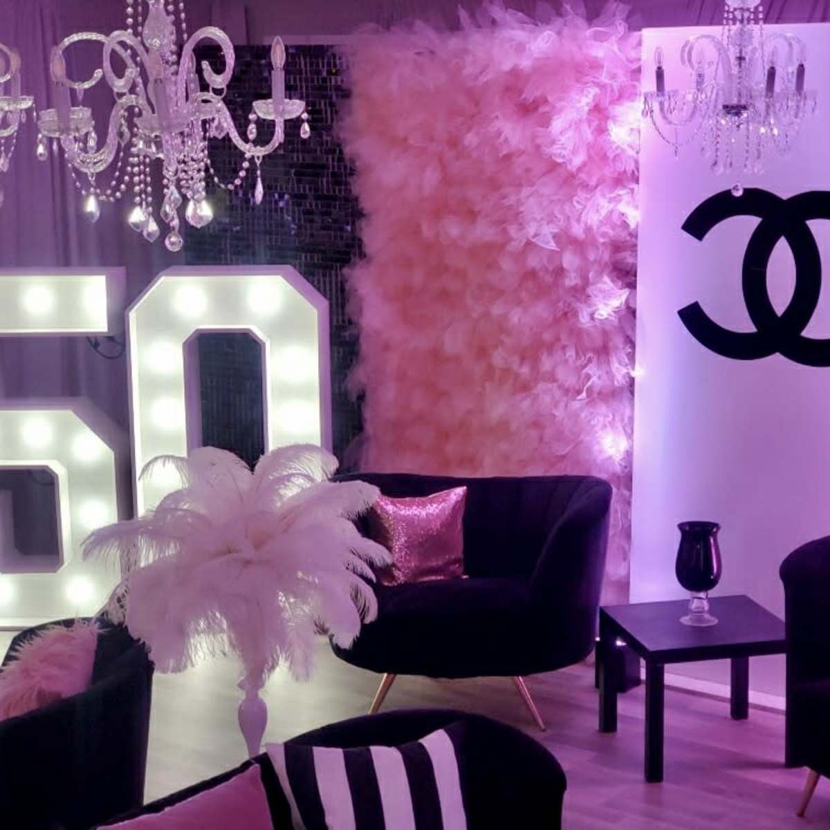 A decorative party setting featuring large illuminated "50" numbers, a Chanel logo backdrop, plush pink decor, and black velvet chairs.