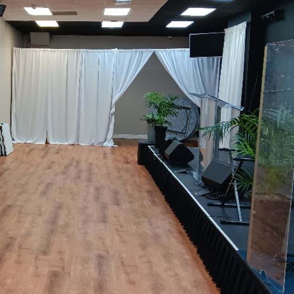 A spacious room with a wooden floor, a stage with musical instruments, and white curtains in the background.