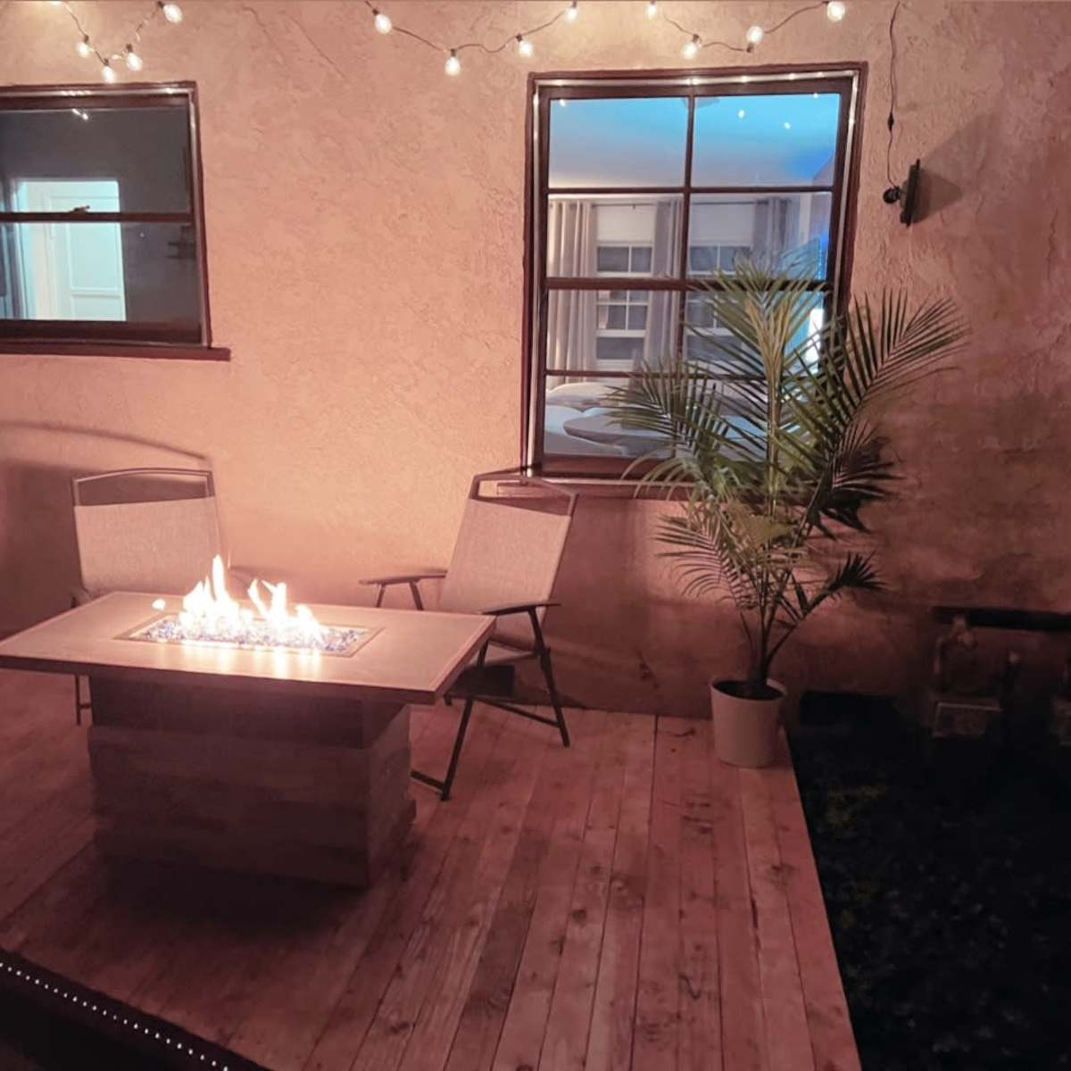 A wooden deck features a fire table surrounded by two chairs and potted plants, with string lights illuminating the area.