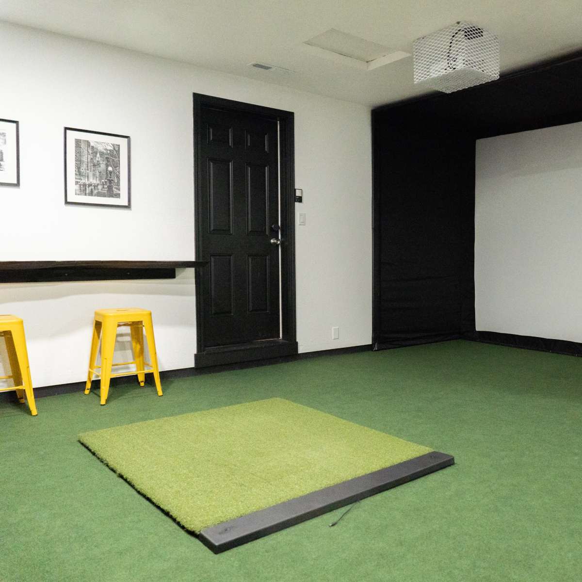An indoor room with a green carpeted floor, featuring a hitting mat in front of a large black projection screen and a door on one side.