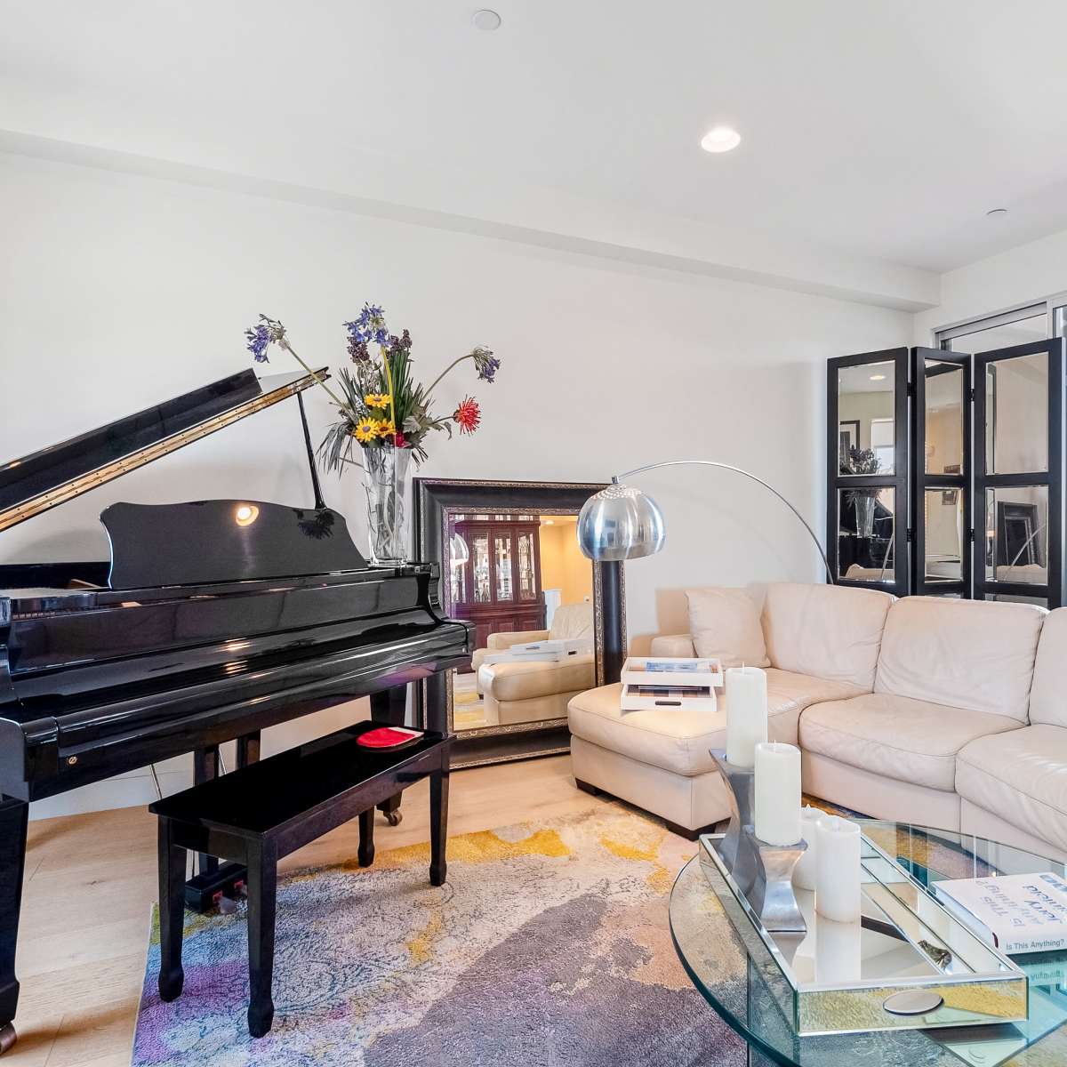 The living room features a grand piano beside a white sectional sofa, a glass coffee table, and large windows with black-frame doors leading outside.