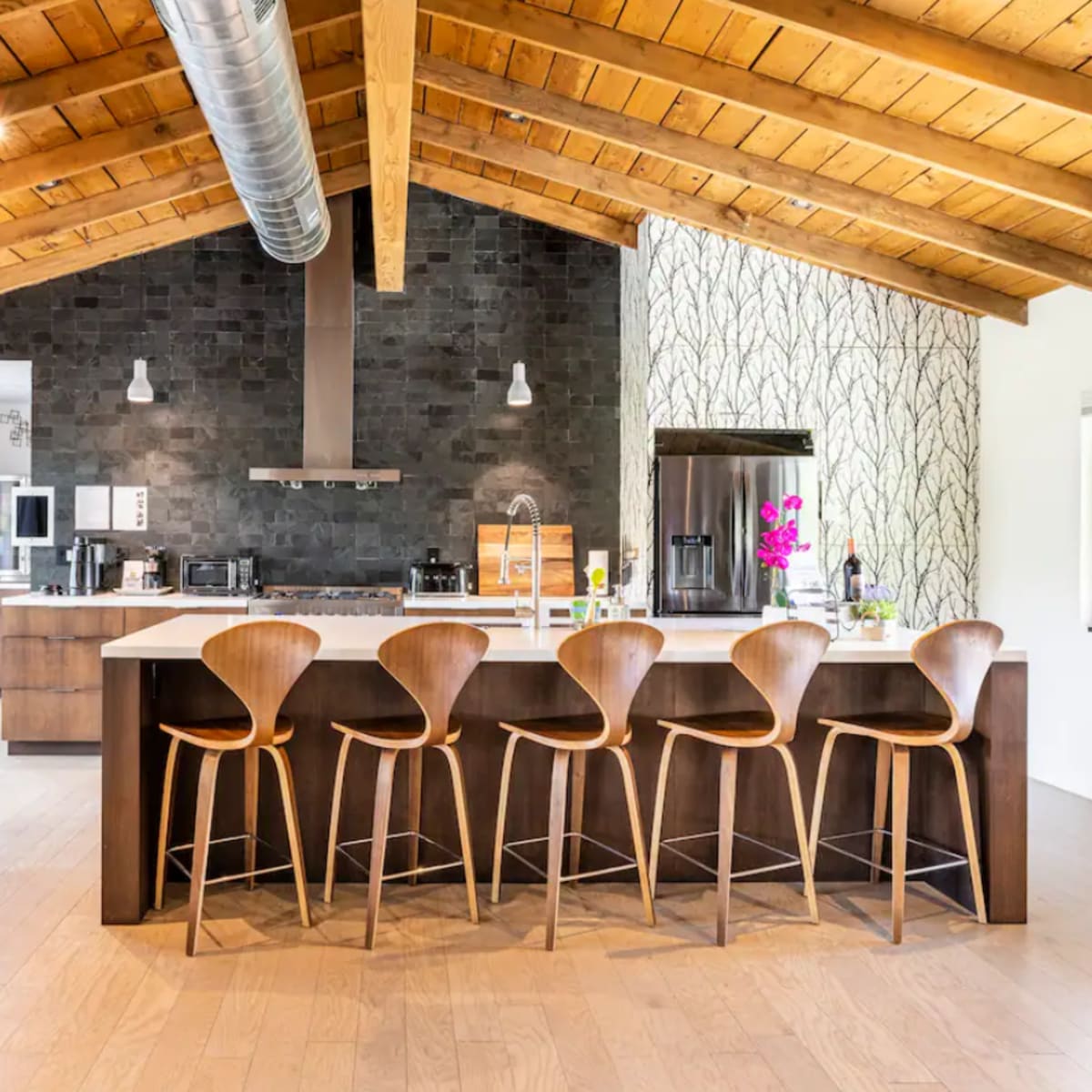 A modern kitchen with a spacious island fitted with wooden bar stools, under a vaulted wooden ceiling and stone accents on the wall.