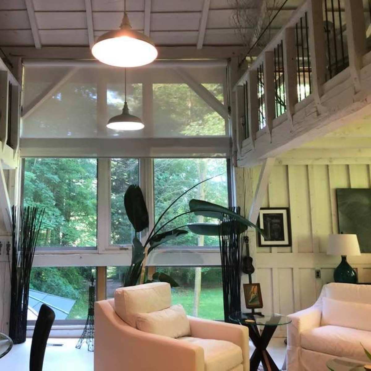 A bright interior of a modern loft-style living space with large windows, white furnishings, and a view of green foliage outside.