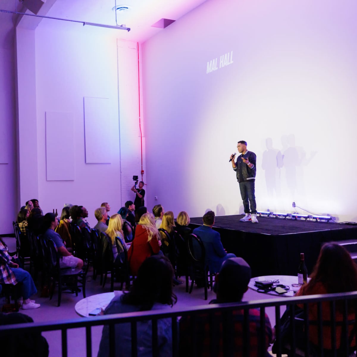 A speaker stands on stage in a modern venue while an audience observes from seated tables.