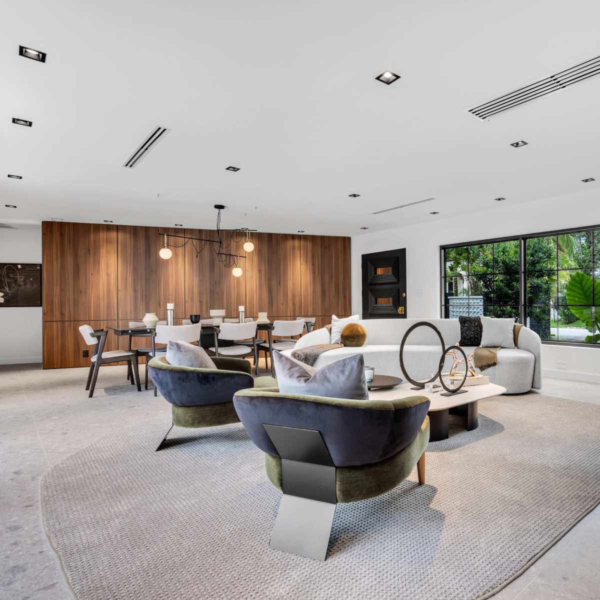 A modern living room features a seating area with chairs and a coffee table, alongside a dining space with a wooden table and chairs, illuminated by pendant lighting.