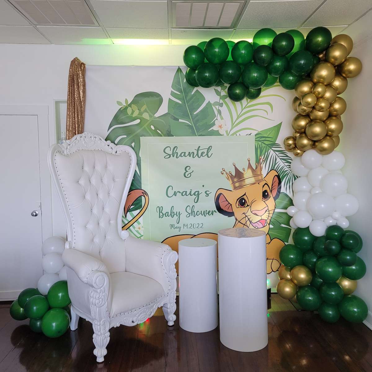 A decorative backdrop for a baby shower featuring a white throne chair, green and gold balloons, and a sign with the names "Shantel & Craig" along with a Lion King theme.