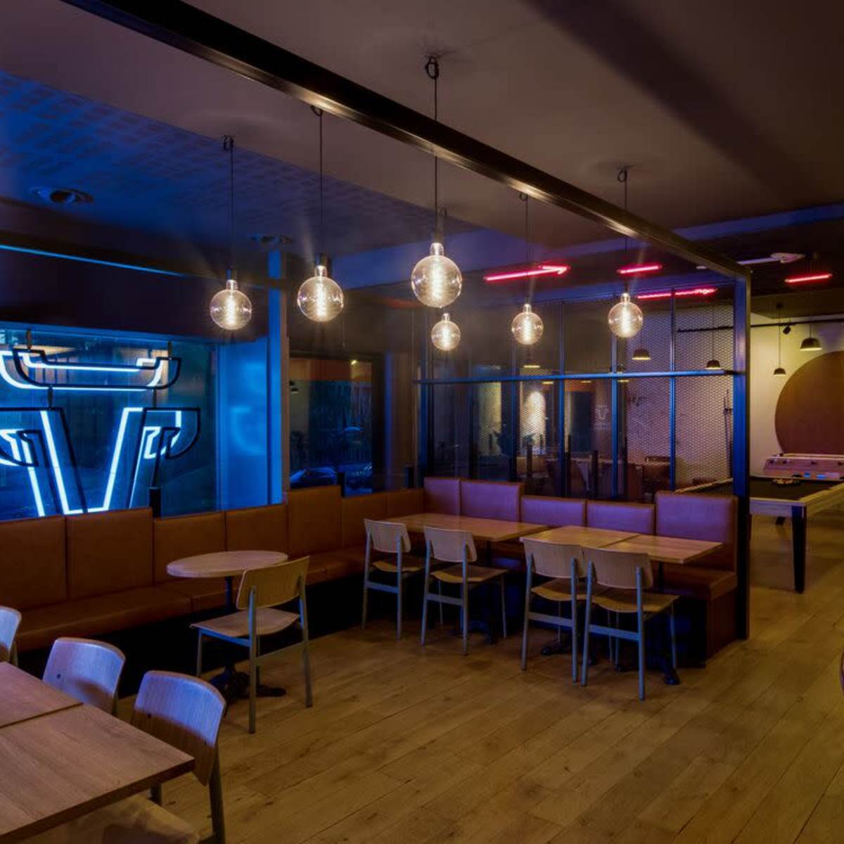 A modern cafe interior with wooden tables and benches, illuminated by pendant lights, featuring a neon sign and a game table in the background.