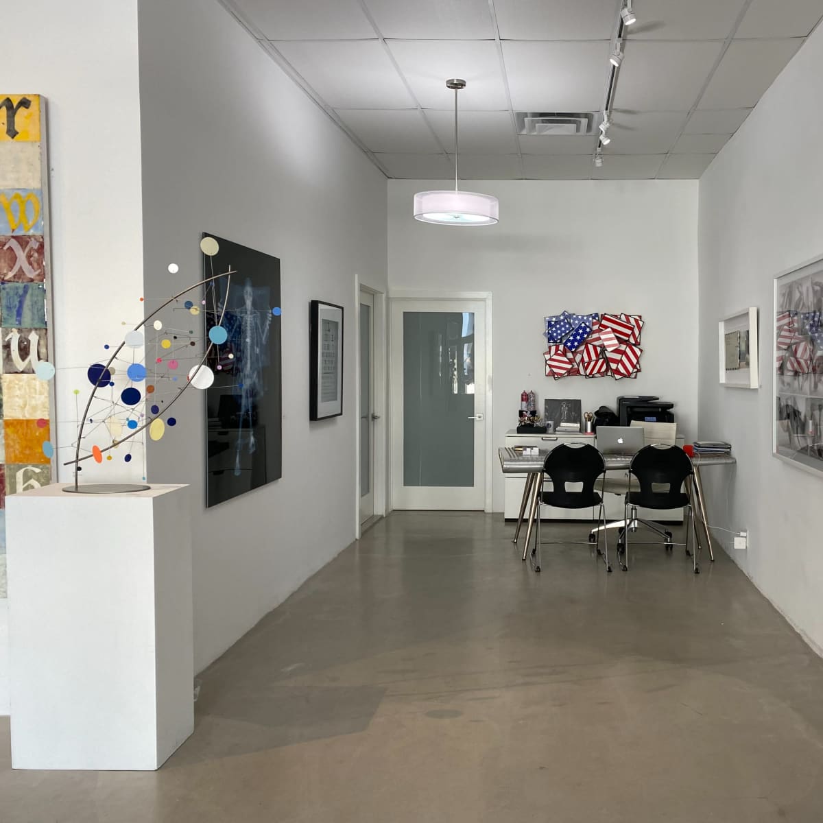 A modern art gallery corridor featuring a reception area with a desk and two chairs, flanked by artworks on the walls and a sculpture in the foreground.