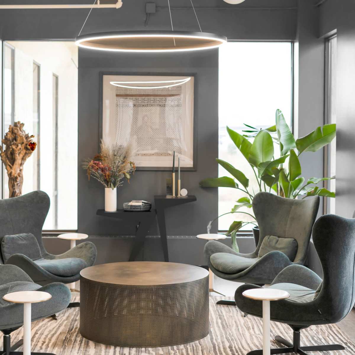 A modern lounge area with four green chairs arranged around a round metal table, alongside large windows and two potted plants.