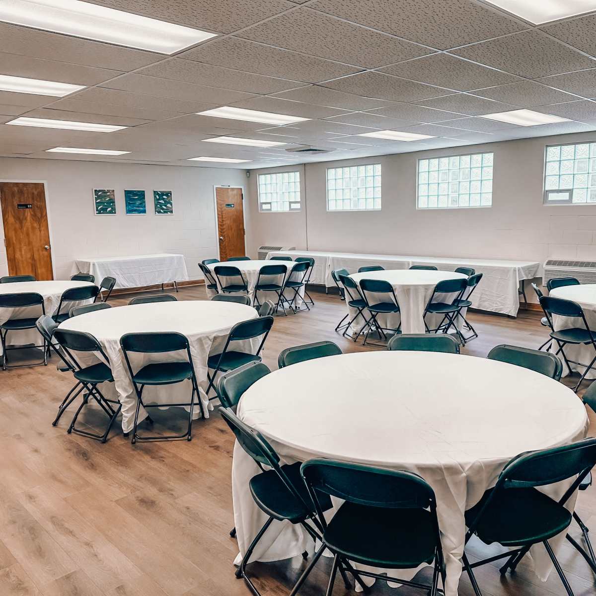A spacious room with multiple round tables covered in white tablecloths and surrounded by green chairs.
