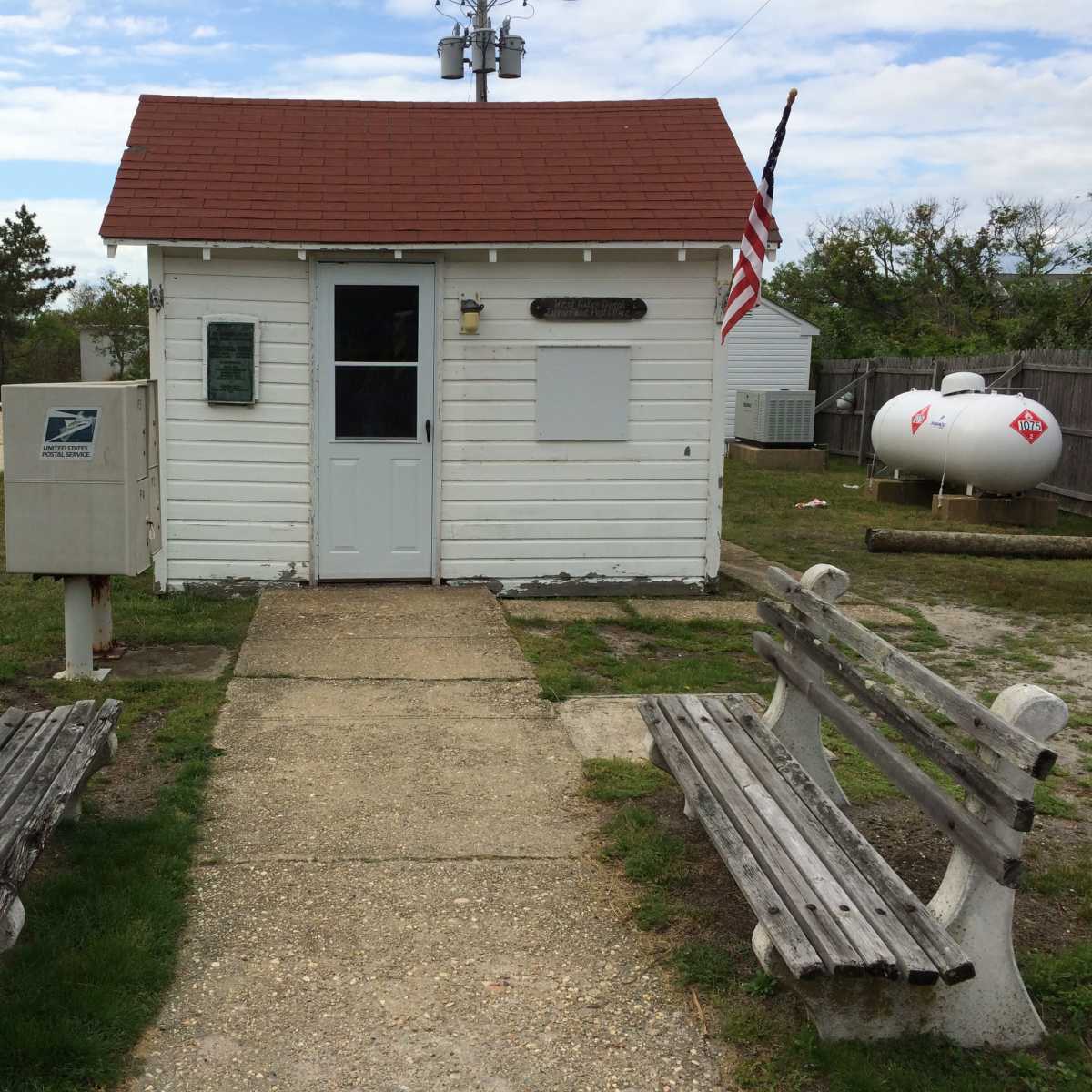 A small white building with a red roof and an American flag stands at the end of a concrete path, flanked by two wooden benches and a mailbox nearby.