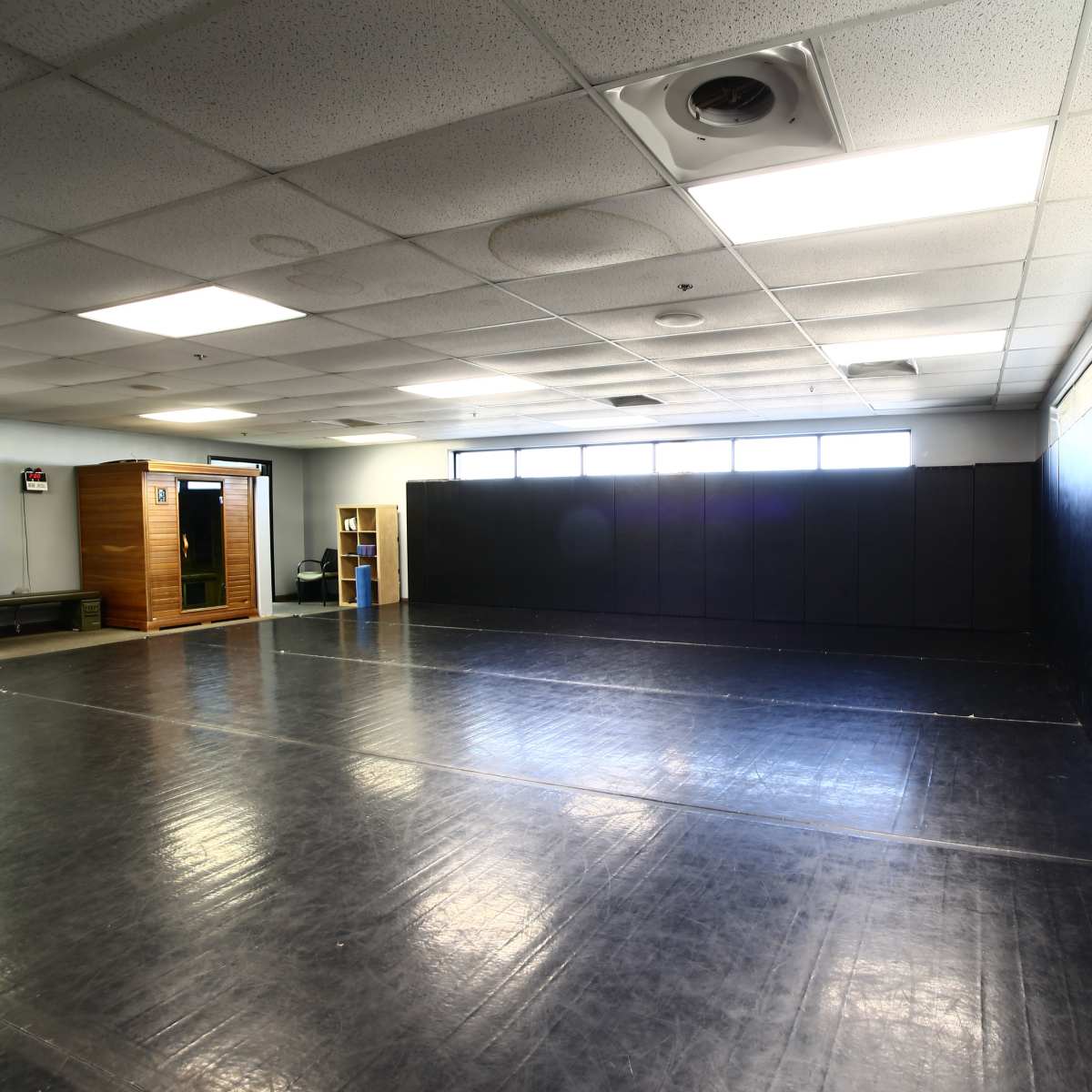 A spacious training room with black mats, large windows, and a wooden structure in one corner.