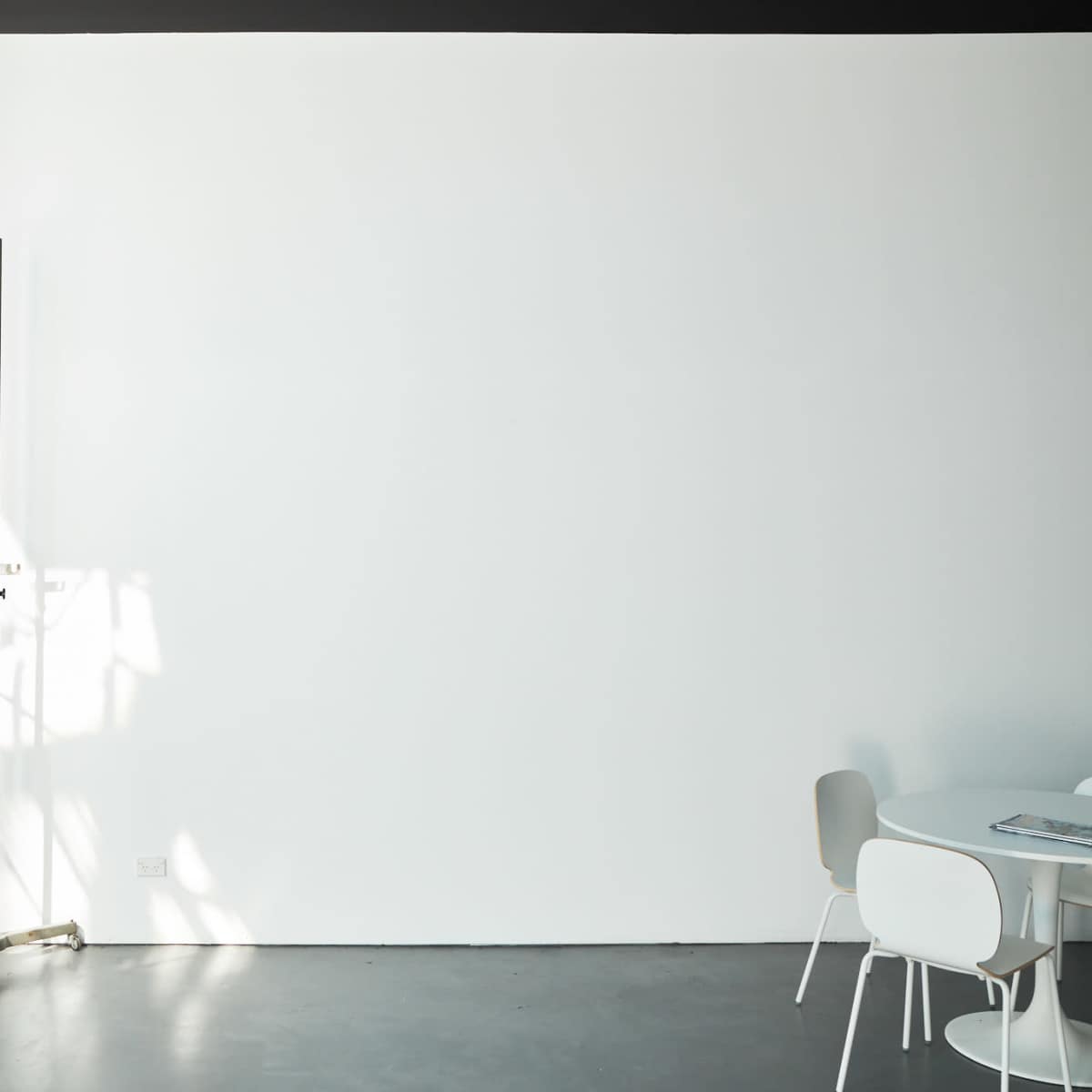 A minimalist room features a blank white wall, a draped curtain, a stand, and a small dining area with simple chairs and a table.