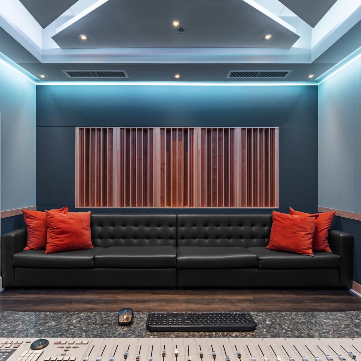 A modern recording studio lounge with a black leather sofa, orange cushions, and a control panel in the foreground.