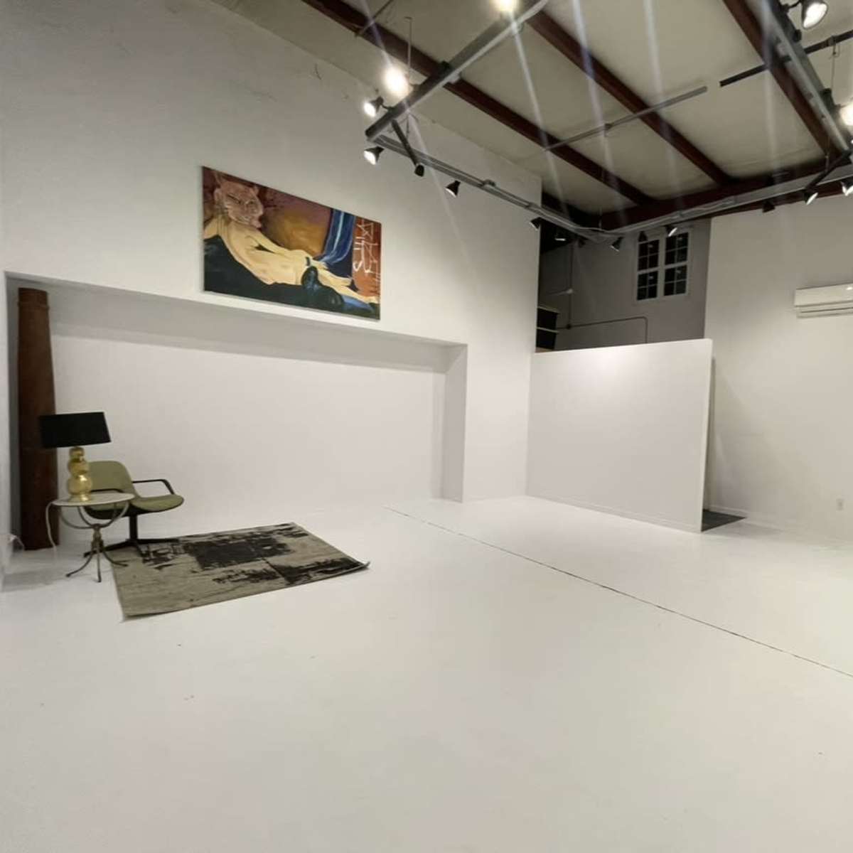 A spacious, empty room with white walls, a wooden beam, and a dark rug on the floor, featuring a painting on the wall and a chair in one corner.