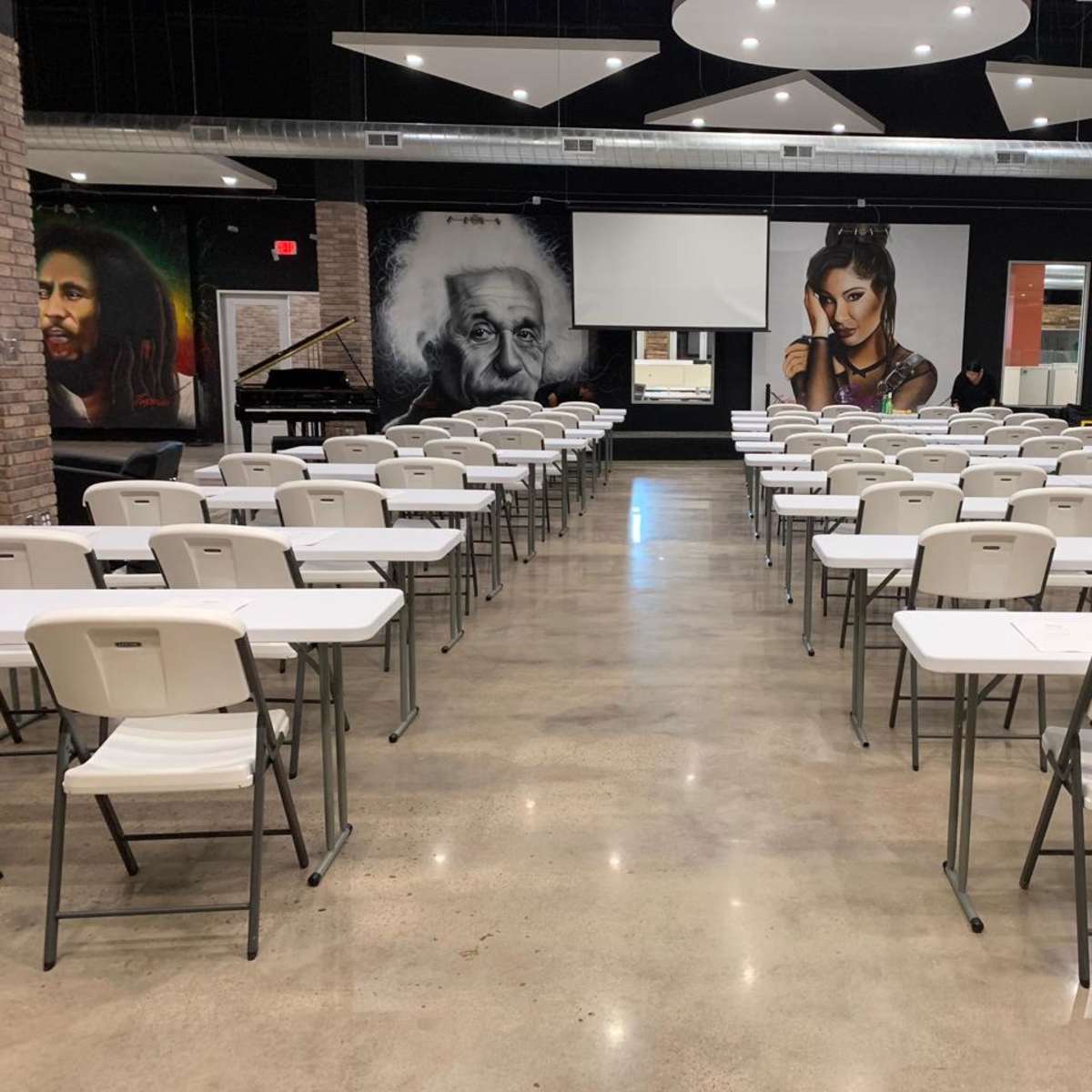A spacious event room with rows of white folding tables and chairs, and large murals of notable figures on the walls.