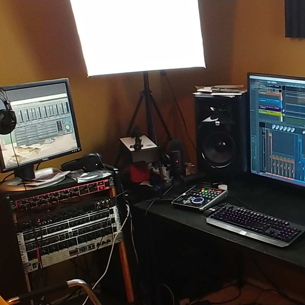 A home studio setup with a large computer monitor displaying audio software, studio monitors on either side, and a second screen beside a microphone, headphones, and recording equipment.