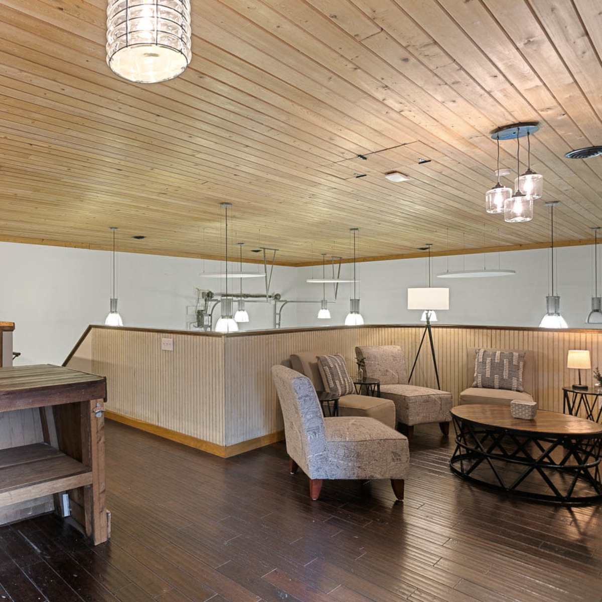 A spacious room with wooden flooring and a ceiling lined with wood panels, featuring a lounge area with plush chairs and small tables, as well as a bar-like wooden counter.