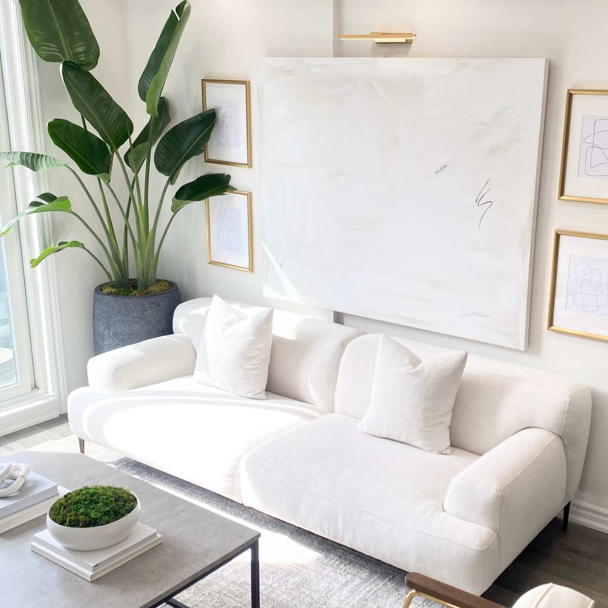 A modern living room features a white sofa with two cushions, a large abstract painting on the wall, framed artworks, and a tall plant beside a black shelving unit.