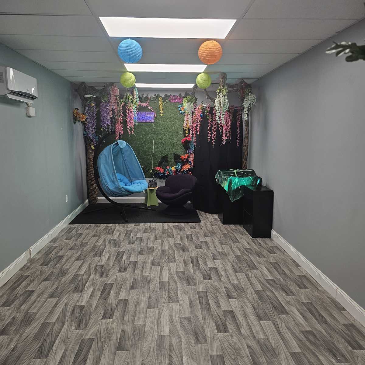 A spacious room features a blue hammock chair, decorative plants, and colorful lanterns hanging from the ceiling, with a green backdrop and wooden flooring.