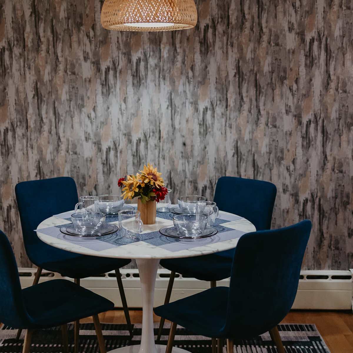 A round dining table with four blue chairs is set with clear glassware and a small vase of flowers, located against a textured wallpaper background.