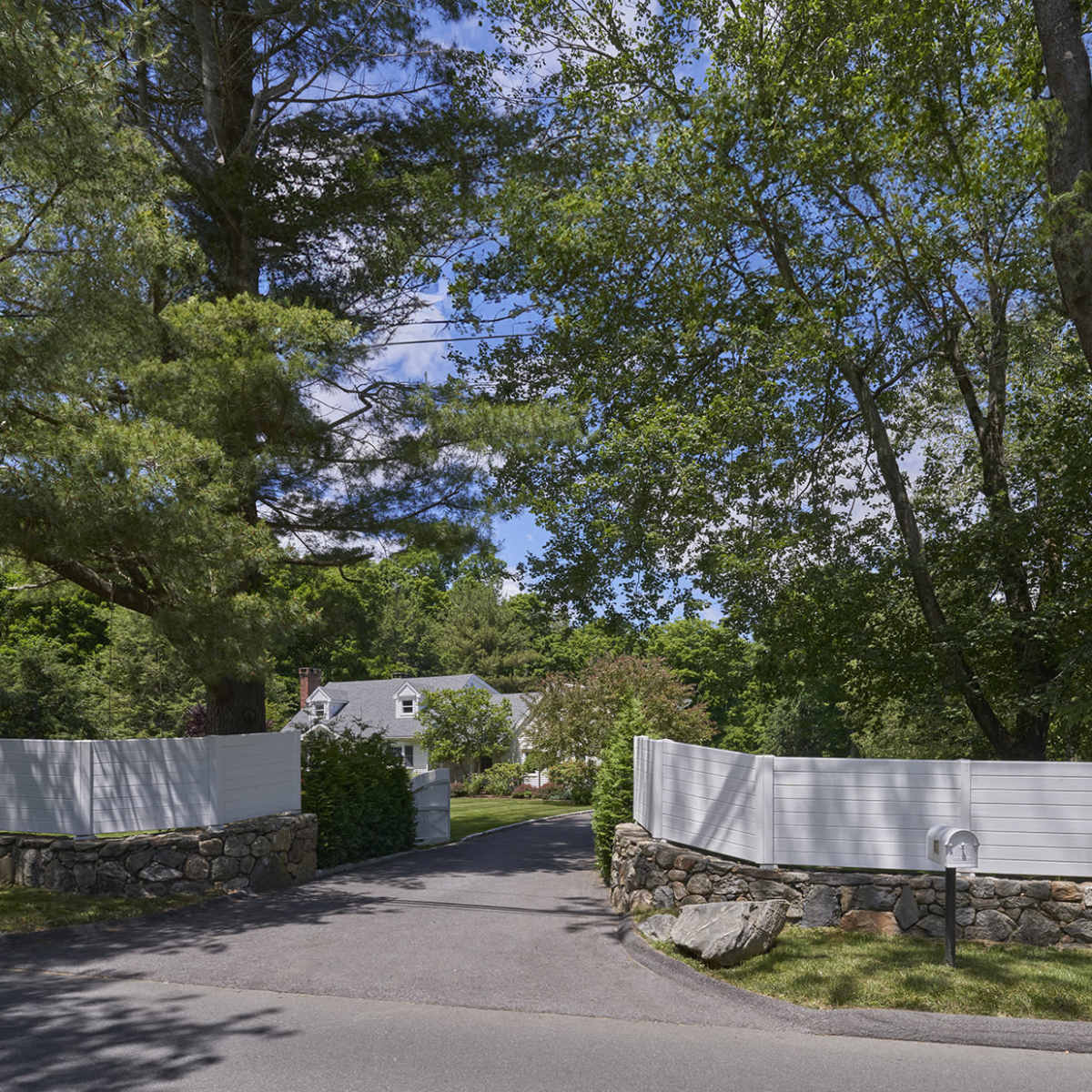 A winding driveway leads through a tree-lined entrance to a home behind white fences and stone walls.