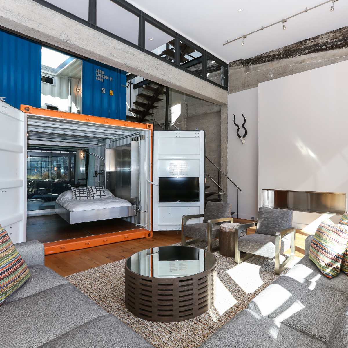 A modern living room featuring a shipping container transformed into a bedroom, with large windows, a sectional sofa, and decorative accents.