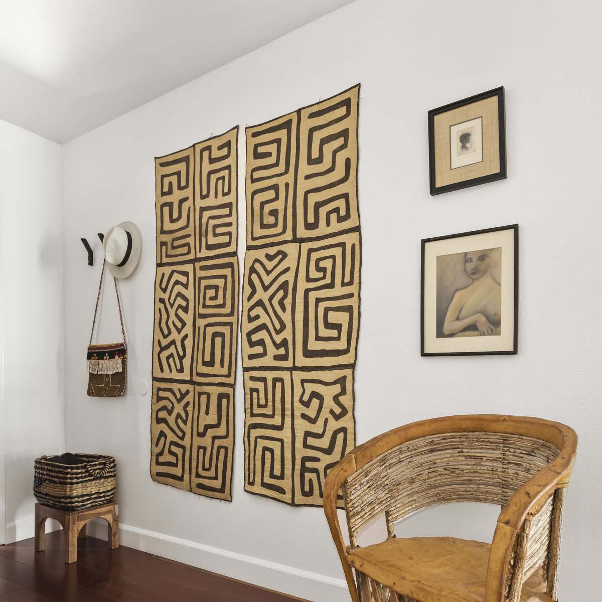A corner of a room features a patterned wall hanging, a wooden chair, and framed artwork, with a hat and a bag hanging nearby.