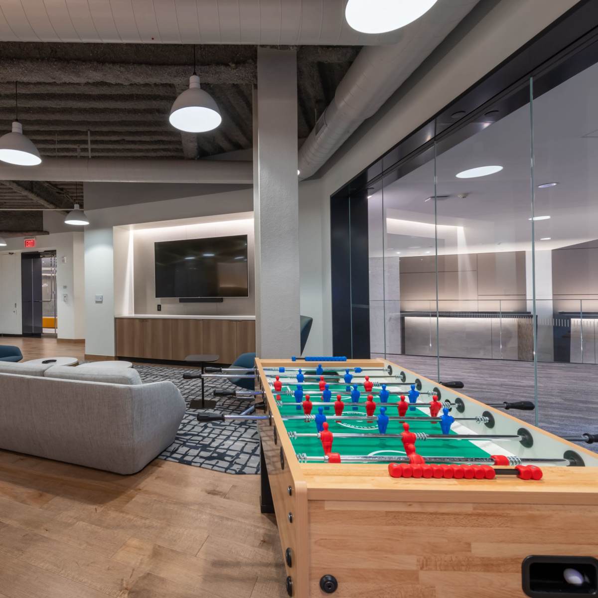 A modern office lounge area featuring a foosball table, a gray sofa, and various seating options under pendant lighting.