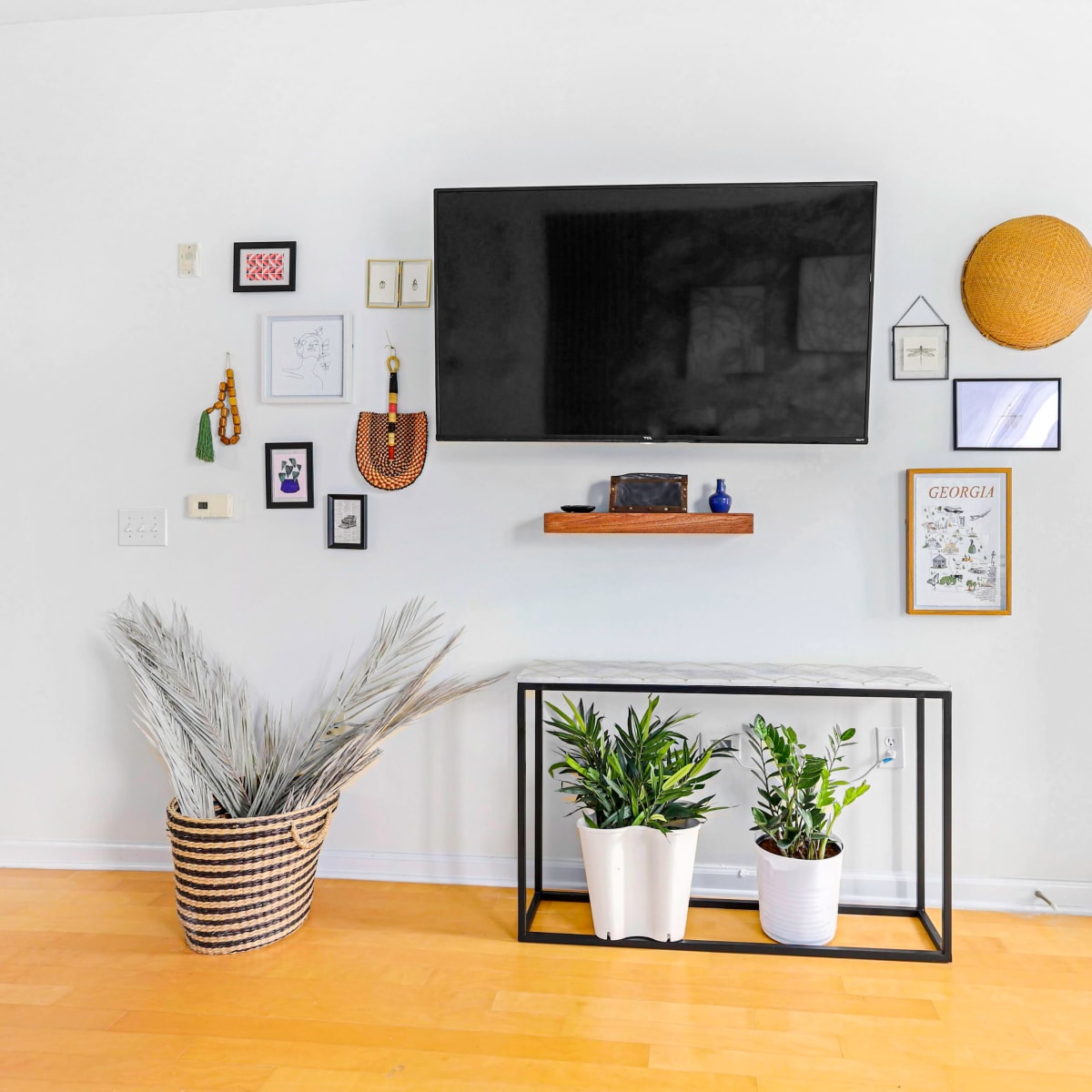 A modern living room wall features a mounted flat-screen TV above a console table, adorned with plants and framed artwork.
