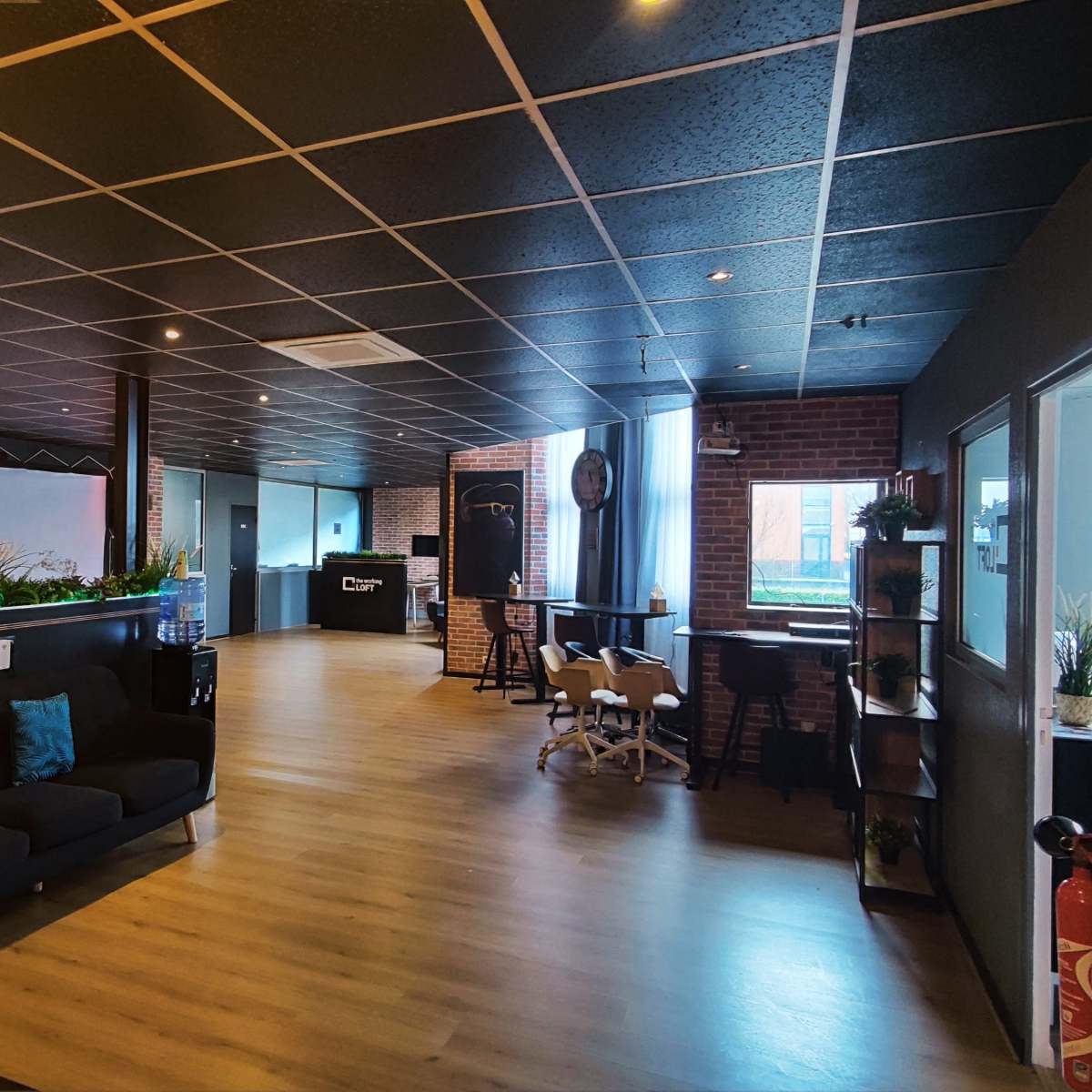 A spacious office interior featuring a lounge area with a black sofa, plants, and modern furnishings, alongside a large open workspace with a polished wooden floor.