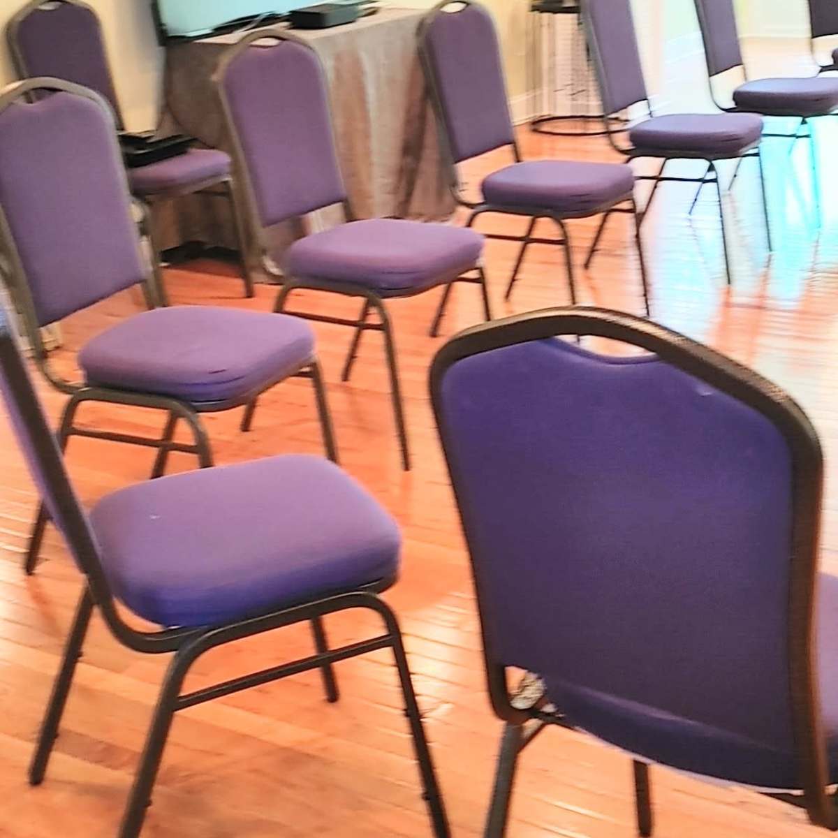 A room with wooden floors contains a circular arrangement of purple chairs and a television display on a stand.