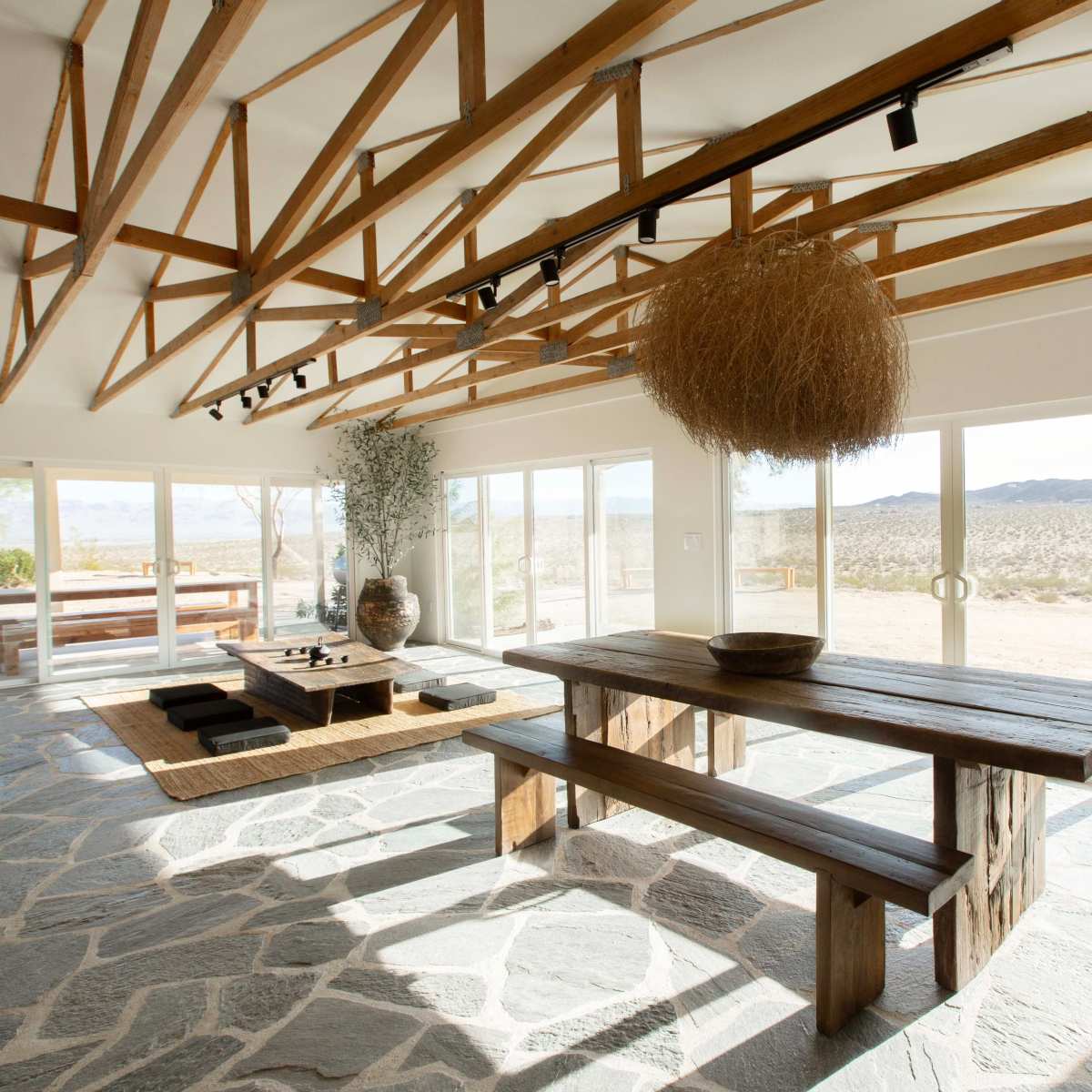 The interior of a spacious room features a wooden table and benches, with large windows showcasing a desert landscape and exposed wooden beams overhead.