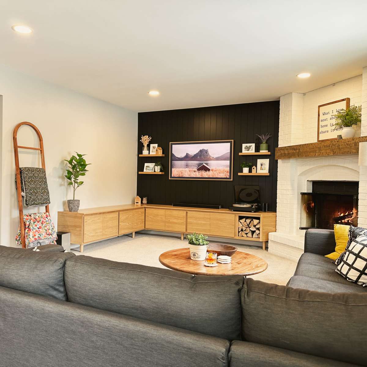 A modern living room with a gray sectional sofa, a circular coffee table, a TV mounted on the wall, and a fireplace surrounded by light-colored brick.