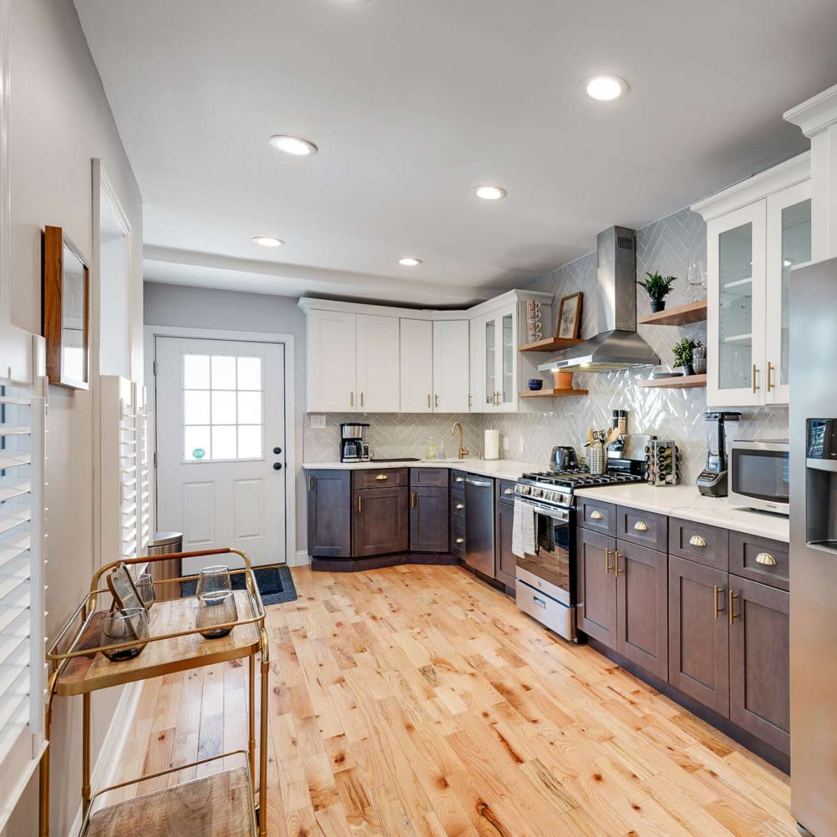 A modern kitchen featuring a stainless steel refrigerator, a gas stove, and a mix of dark and light cabinetry, with wooden flooring and a door leading outside.